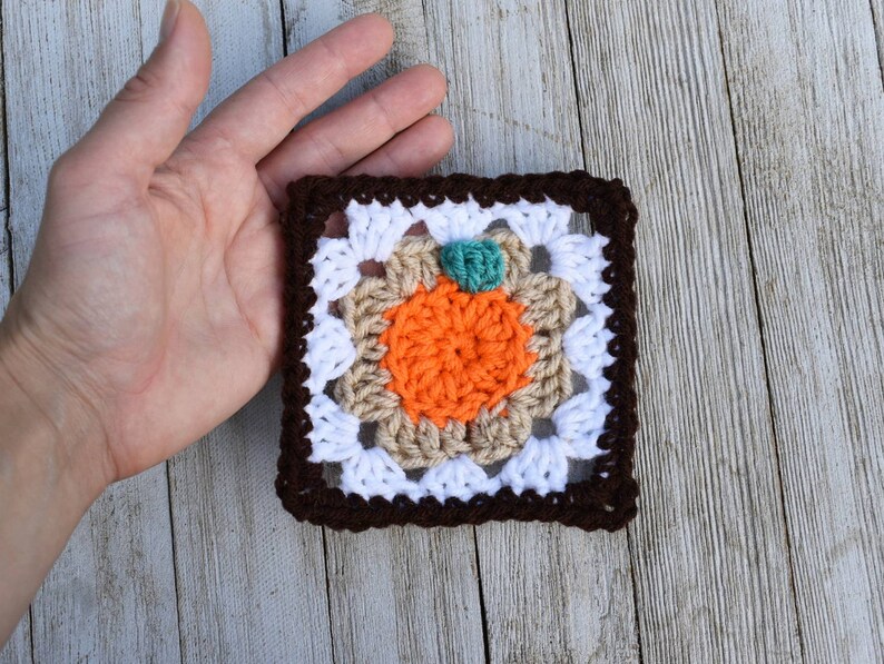 May include: A crocheted square with a brown border, white center, and an orange pumpkin in the middle. The pumpkin has a green stem.