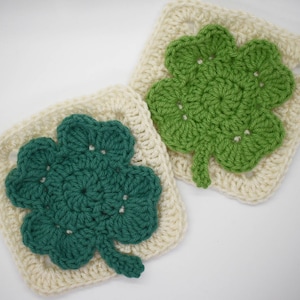 May include: Two crocheted squares with a four-leaf clover design. One square is white with a green clover, and the other is white with a teal clover.