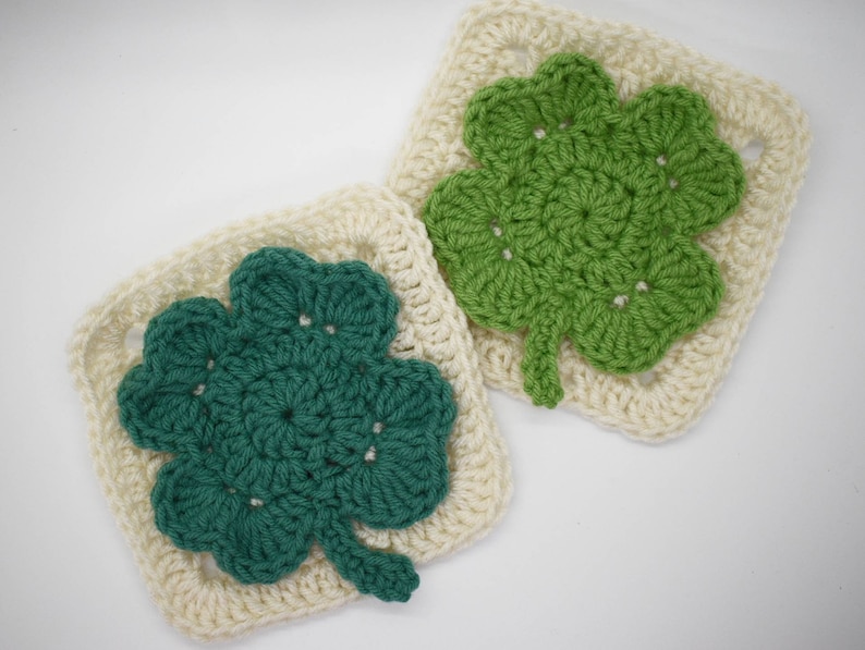 May include: Two crocheted squares with a four-leaf clover design. One square is white with a green clover, and the other is white with a teal clover.