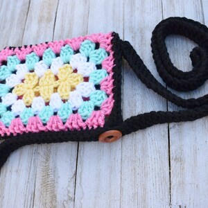 May include: A small crocheted bag with a black strap and a wooden button closure. The bag is made with a granny square pattern in pink, white, yellow, and turquoise yarn.