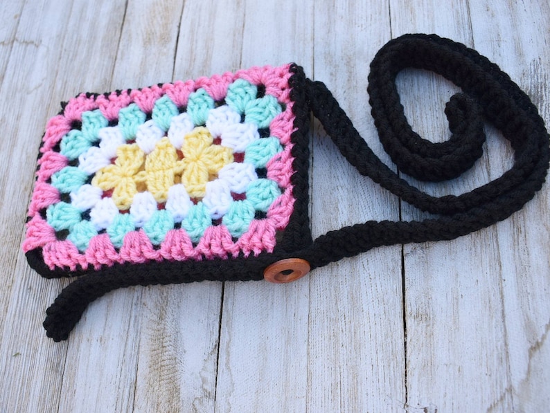 May include: A small crocheted bag with a black strap and a wooden button closure. The bag is made with a granny square pattern in pink, white, yellow, and turquoise yarn.