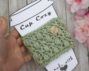 Könnte beinhalten: Ein grüner gehäkelter Becherwärmer mit einem Knopf und dem Text "Cup Cozy" und "made with" mit einem Herzen.