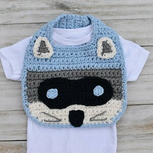 May include: A blue, gray, and white crocheted baby bib with a raccoon face design. The bib has two black eyes, a black nose, and white whiskers.