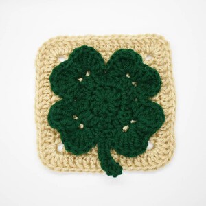 May include: A crocheted square with a green four-leaf clover in the center. The square is beige and the clover is green.