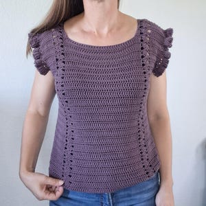 May include: A mauve crochet top featuring a wide scoop neckline, short ruffled sleeves, and vertical openwork stitch patterns running down the front sides. The garment is constructed with a dense, textured yarn in a solid muted purple color.
