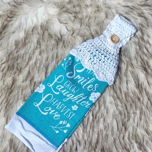 Puede incluir: Una toalla de cocina de color turquesa con texto blanco que dice "That smiles grow laughter harvest love". La toalla tiene un lazo de crochet blanco con un botón de madera.