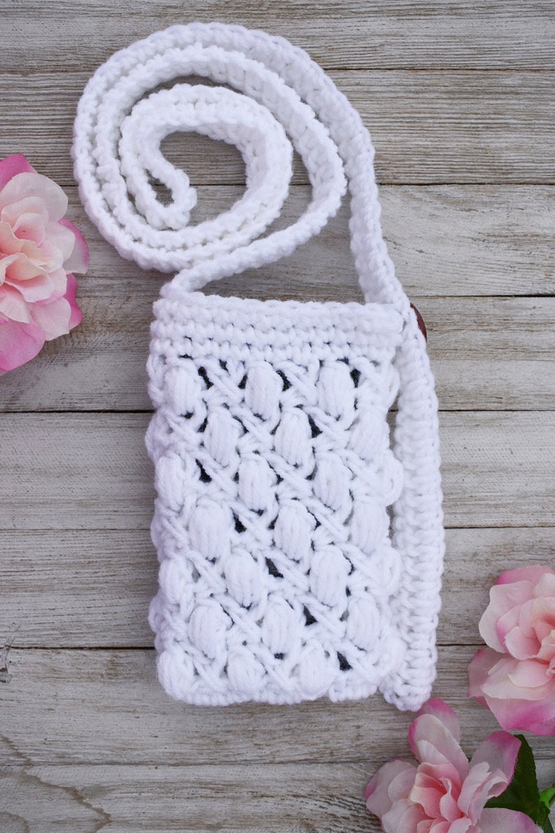 May include: White crocheted crossbody bag with a textured, puffy design. The bag has a long strap and is made with a white yarn.