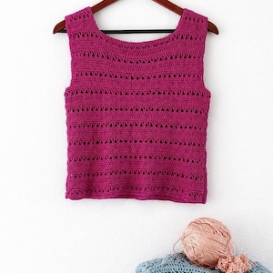 May include: A magenta crochet vest top hangs on a wooden hanger. The top has a scoop neckline and horizontal rows of openwork. A ball of peach-coloured yarn and folded crochet items in blue and green are below.