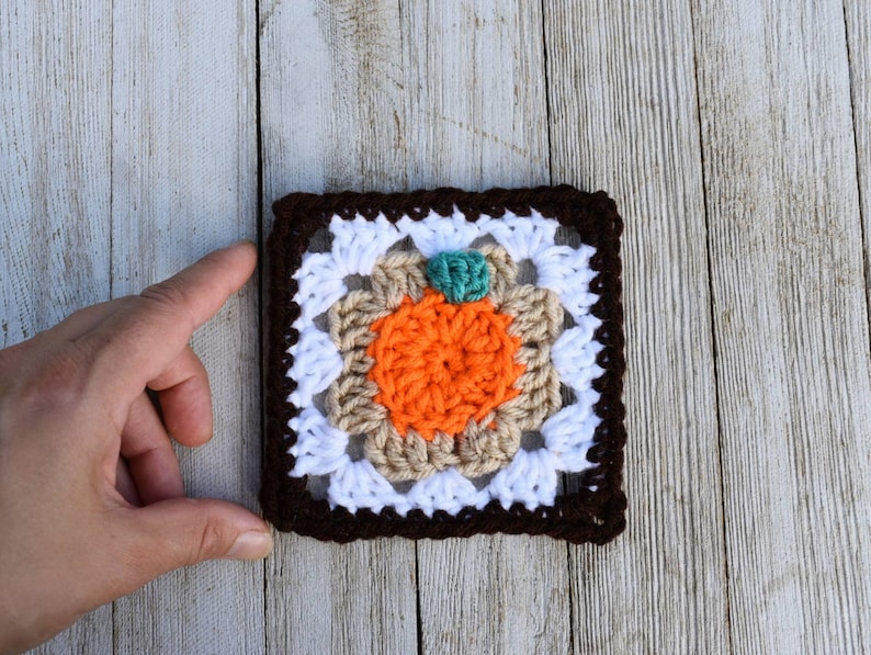 May include: A crocheted square with a brown border, white center, and an orange pumpkin in the middle. The pumpkin has a green stem.