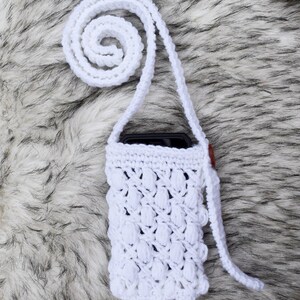 May include: White crocheted phone case with a long strap. The case has a textured design and is shown on a white faux fur background.