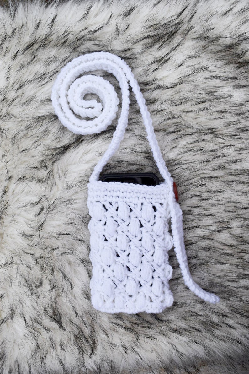 May include: White crocheted phone case with a long strap. The case has a textured design and is shown on a white faux fur background.