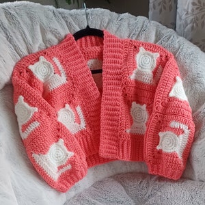 Könnte beinhalten: Eine rosa gehäkelte Strickjacke mit weißen Katzenapplikationen. Die Strickjacke hat einen V-Ausschnitt und lange Ärmel.