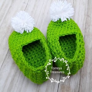 May include: A pair of green crocheted baby booties with white pom-poms on top. The booties are made with a textured stitch and have a simple, classic design.