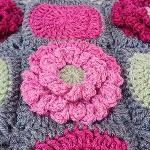 May include: A close-up of a crocheted blanket with a variety of pink and green flowers. The blanket is made of a soft, textured yarn and has a gray background.