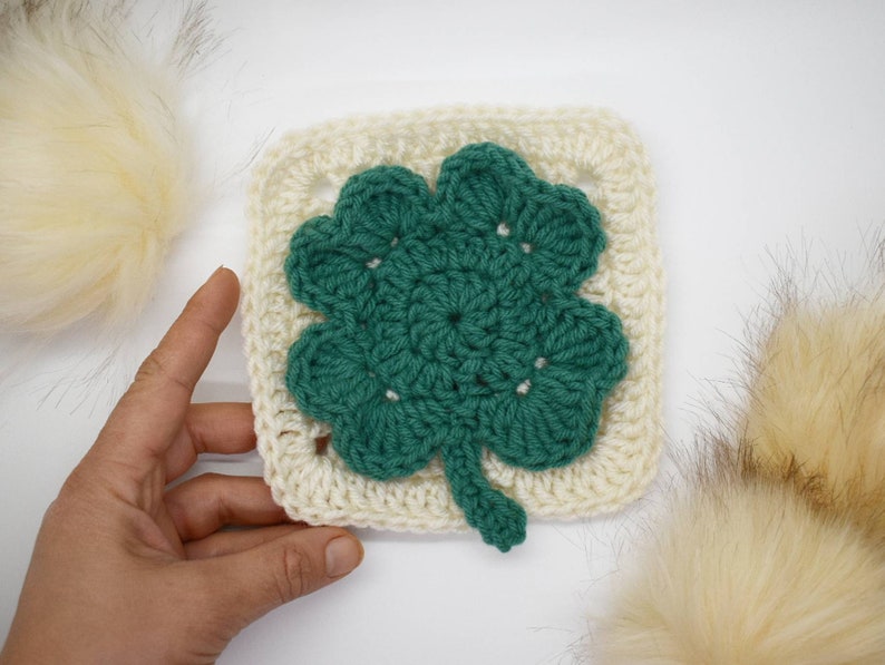 May include: A crocheted square with a green four-leaf clover design in the center. The square is made with white yarn and the clover is made with green yarn.