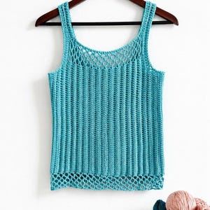 May include: A turquoise crochet tank top with a ribbed design, hanging on a wooden hanger. The top features a mesh neckline and a decorative bottom edge. A ball of yarn and folded fabric are visible below.