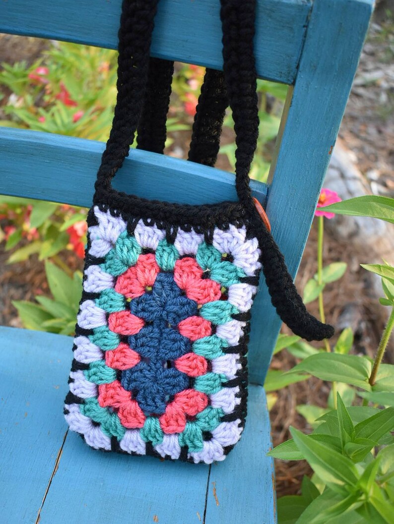 May include: A colourful crocheted granny square bag with a black strap. The bag is hanging on a blue wooden chair.