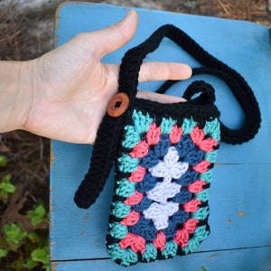 May include: A black crocheted crossbody bag with a colourful granny square design in shades of pink, teal, and purple. The bag has a black strap and a wooden button.