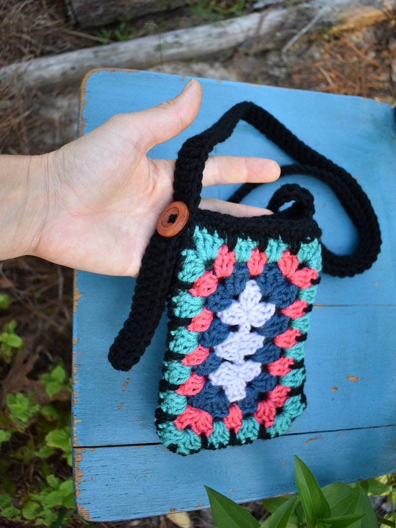 May include: A black crocheted crossbody bag with a colourful granny square design in shades of pink, teal, and purple. The bag has a black strap and a wooden button.
