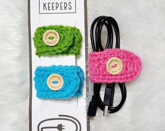 May include: Three crocheted cord keepers with wooden buttons that say "Handmade with love". The cord keepers are in green, pink, and blue.
