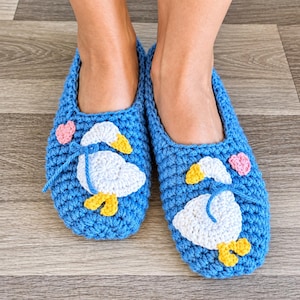 May include: A pair of bright blue crochet slippers featuring white goose appliqués with yellow beaks and feet. Each slipper is decorated with a small pink heart accent. The slippers are crafted with a textured yarn stitch and have a rounded toe design.