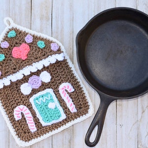 Puede incluir: Un posavasos de ganchillo en forma de casa de jengibre con ribete blanco, detalles de bastones de caramelo y decoraciones coloridas. El posavasos está al lado de una sartén de hierro fundido negra.