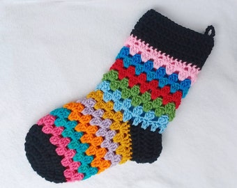 Może przedstawiać: Ręcznie robiona, szydełkowa skarpeta bożonarodzeniowa z żywym wzorem granny square. Skarpeta ma czarny mankiet i palce, z tęczą kolorów, w tym różowym, niebieskim, zielonym, czerwonym, pomarańczowym i żółtym. Do zawieszenia przymocowana jest pętla.