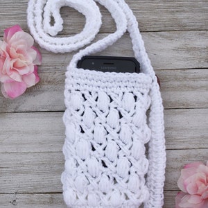 May include: White crocheted phone case with a long strap. The case has a textured design and is holding a black Samsung smartphone.