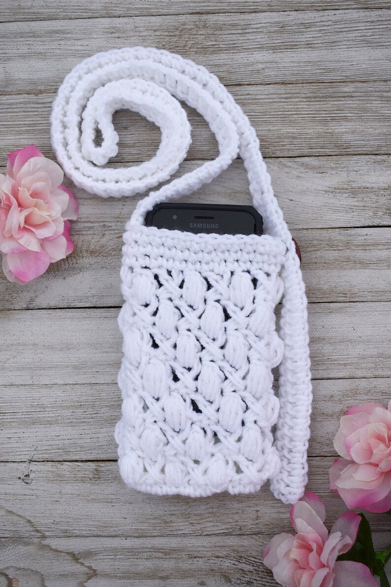 May include: White crocheted phone case with a long strap. The case has a textured design and is holding a black Samsung smartphone.