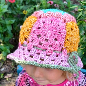 May include: A young child wearing a pink, orange, and blue crocheted sun hat with a flower design. The hat has a wide brim and is made with a soft, textured yarn.