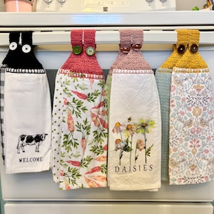 May include: Four kitchen towels hanging on a rack. The towels have different designs, including a cow with the word "WELCOME" underneath, carrots, daisies, and a floral pattern. The towels have crocheted loops with buttons at the top.