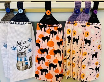 Puede incluir: Un conjunto de paños de cocina colgantes con la parte superior de ganchillo y cierres de botón. Las toallas presentan varios diseños, incluyendo un gato con bufanda, calabazas y gatos negros, y patrones de Halloween. Las toallas son blancas, rosas y naranjas.