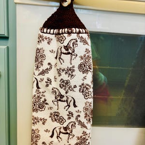 Op de afbeelding: Een hangende keukendoek met een witte stoffen basis en een herhalende bruine print van paarden en bloemen. Het bovenste gedeelte is gemaakt van donkerbruin gehaakt garen en bevat een enkele lichtgekleurde knoop voor bevestiging.