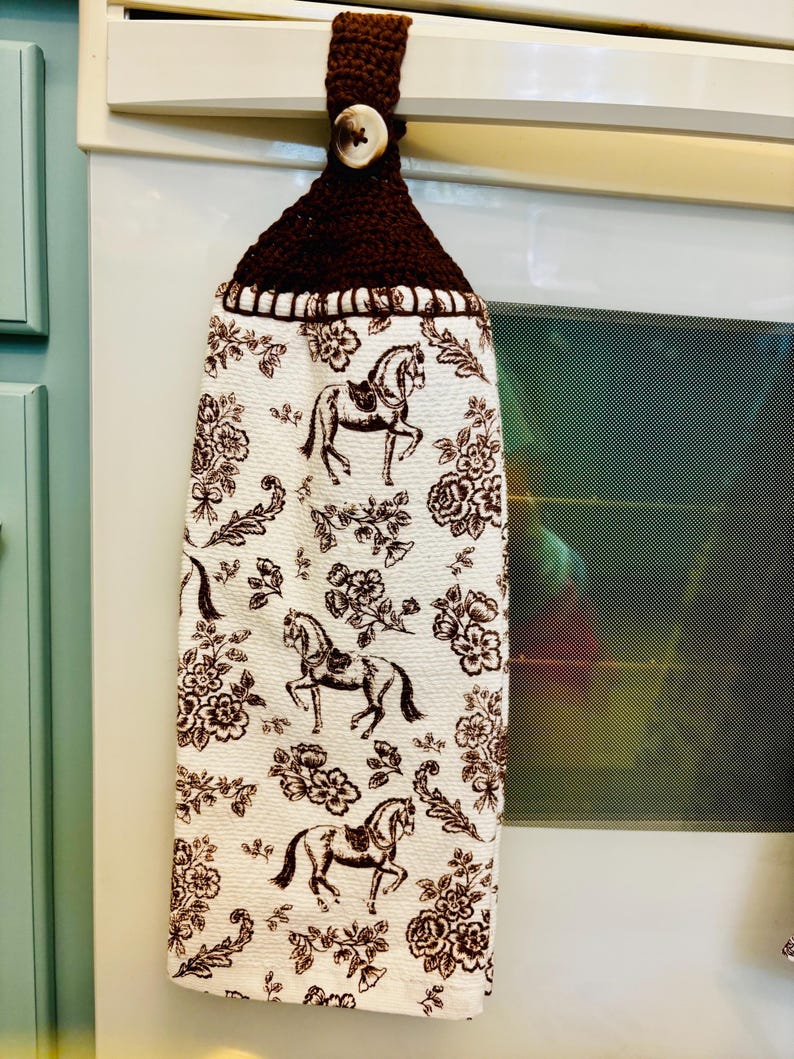 Op de afbeelding: Een hangende keukendoek met een witte stoffen basis en een herhalende bruine print van paarden en bloemen. Het bovenste gedeelte is gemaakt van donkerbruin gehaakt garen en bevat een enkele lichtgekleurde knoop voor bevestiging.