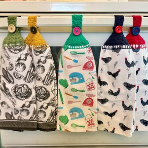 Op de afbeelding: Vijf hangende keukendoeken met gehaakte bovenkanten in olijf, mosterd, groen, marineblauw en rood, elk vastgezet met een bijpassende knoop. De doeken hebben diverse prints, waaronder zwart-witte groenten, kleurrijke keukengerei en zwarte hanen.