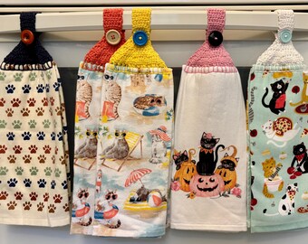 Puede incluir: Cinco paños de cocina colgantes con la parte superior de ganchillo y cierres de botón. Las toallas presentan varios diseños con temas de gatos, incluyendo huellas de patas, escenas de playa y motivos de Halloween. Las toallas están colgadas en un electrodoméstico blanco.