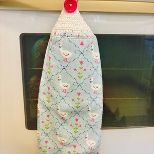 Op de afbeelding: Een hangende keukendoek van lichtblauwe stof met een herhalend patroon van witte ganzen en roze bloemen op een donkerblauw ruitpatroon. De bovenkant is afgewerkt met een witte gehaakte lus die vastzit met een felroze knoop.