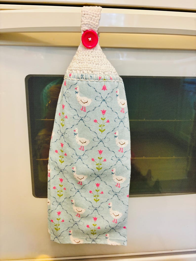 Op de afbeelding: Een hangende keukendoek van lichtblauwe stof met een herhalend patroon van witte ganzen en roze bloemen op een donkerblauw ruitpatroon. De bovenkant is afgewerkt met een witte gehaakte lus die vastzit met een felroze knoop.