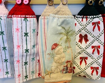Puede incluir: Una colección de paños de cocina decorativos con la parte superior de ganchillo y cierres de botón. Las toallas presentan varios patrones, incluyendo rayas, lazos y una escena de playa con un muñeco de nieve. La paleta de colores incluye rojo, verde, blanco y azul.