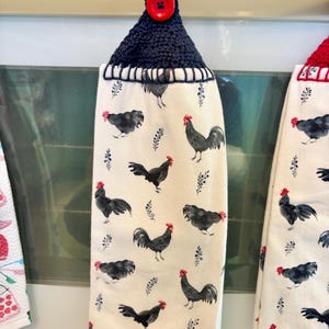 Op de afbeelding: Een hangende keukendoek van cr&egrave;mekleurige stof met een herhalend patroon van zwarte hanen met rode kammen. De doek is aan de bovenkant voorzien van een donker marineblauw gehaakt handvat, vastgezet met een enkele, ronde, felrode knoop.