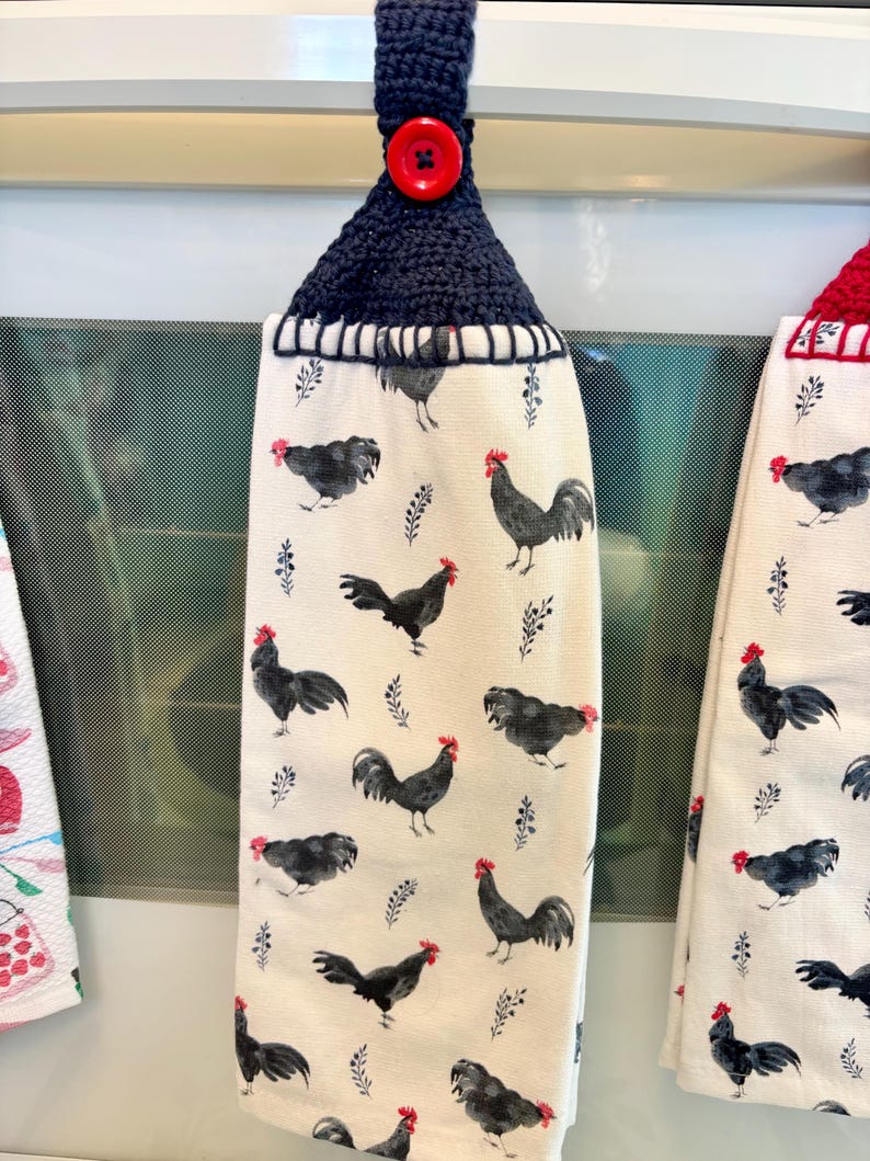 Op de afbeelding: Een hangende keukendoek van cr&egrave;mekleurige stof met een herhalend patroon van zwarte hanen met rode kammen. De doek is aan de bovenkant voorzien van een donker marineblauw gehaakt handvat, vastgezet met een enkele, ronde, felrode knoop.