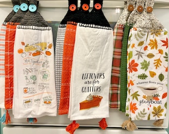 Puede incluir: Una colección de paños de cocina colgantes con la parte superior de ganchillo y botones decorativos. Las toallas presentan diseños con temas de otoño, incluyendo recetas de pastel de calabaza y la frase "Leftovers are for Quitters."