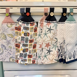 May include: Five kitchen towels hanging on a rack. The towels are made of white fabric with different patterns. The patterns include fish, seashells, and starfish. The towels are all trimmed with black and blue crocheted edging.