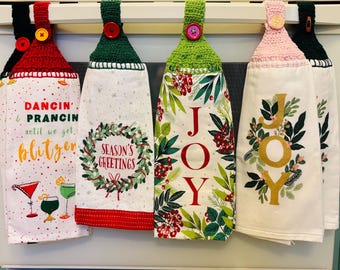 Puede incluir: Un juego de cinco paños de cocina con temática navideña colgados de una barra de metal. Cada paño presenta un diseño único con texto y gráficos festivos, incluyendo "Dancin' & Prancin'", "Season's Greetings" y "JOY". Los paños tienen la parte superior de ganchillo con botones.