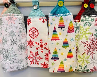 Puede incluir: Cuatro paños de cocina decorativos con la parte superior de ganchillo y cierres de botón. Los paños presentan diseños festivos, incluyendo copos de nieve y árboles de Navidad coloridos. Los paños están colgados de una barra de metal, mostrando una variedad de patrones y colores temáticos de vacaciones.