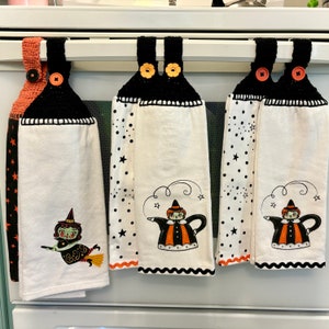 May include: Three Halloween-themed kitchen towels hanging on a white bar. The towels have black crochet tops with decorative buttons. Two towels feature a witch teapot design, and one has a witch on a broom. The towels are white with black polka dots and stars.