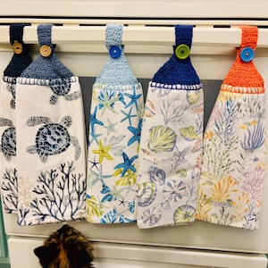 May include: Five hanging kitchen towels with crochet tops and button closures. The towels feature sea-themed patterns, including turtles, starfish, and seashells, in shades of blue, green, and orange. The towels are hanging on a white bar.