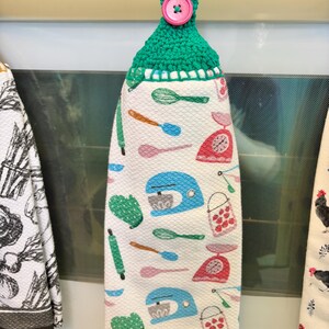 Op de afbeelding: Een hangende keukendoek met een groene gehaakte bovenkant en een roze knoop. De witte doek heeft een herhalend patroon van blauwe keukenmixers, rode keukenweegschalen, groene ovenwanten, groene gardes en roze en blauwe lepels en spatels.