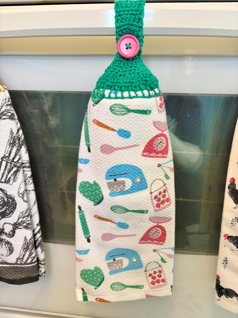 Op de afbeelding: Een hangende keukendoek met een groene gehaakte bovenkant en een roze knoop. De witte doek heeft een herhalend patroon van blauwe keukenmixers, rode keukenweegschalen, groene ovenwanten, groene gardes en roze en blauwe lepels en spatels.
