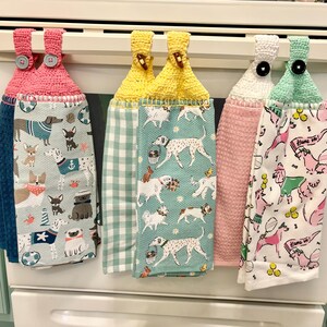 May include: Four kitchen towels with crocheted loops hang on a rack. Each towel features a different pattern of dogs and a different color scheme. The towels are pink, yellow, white, and green.